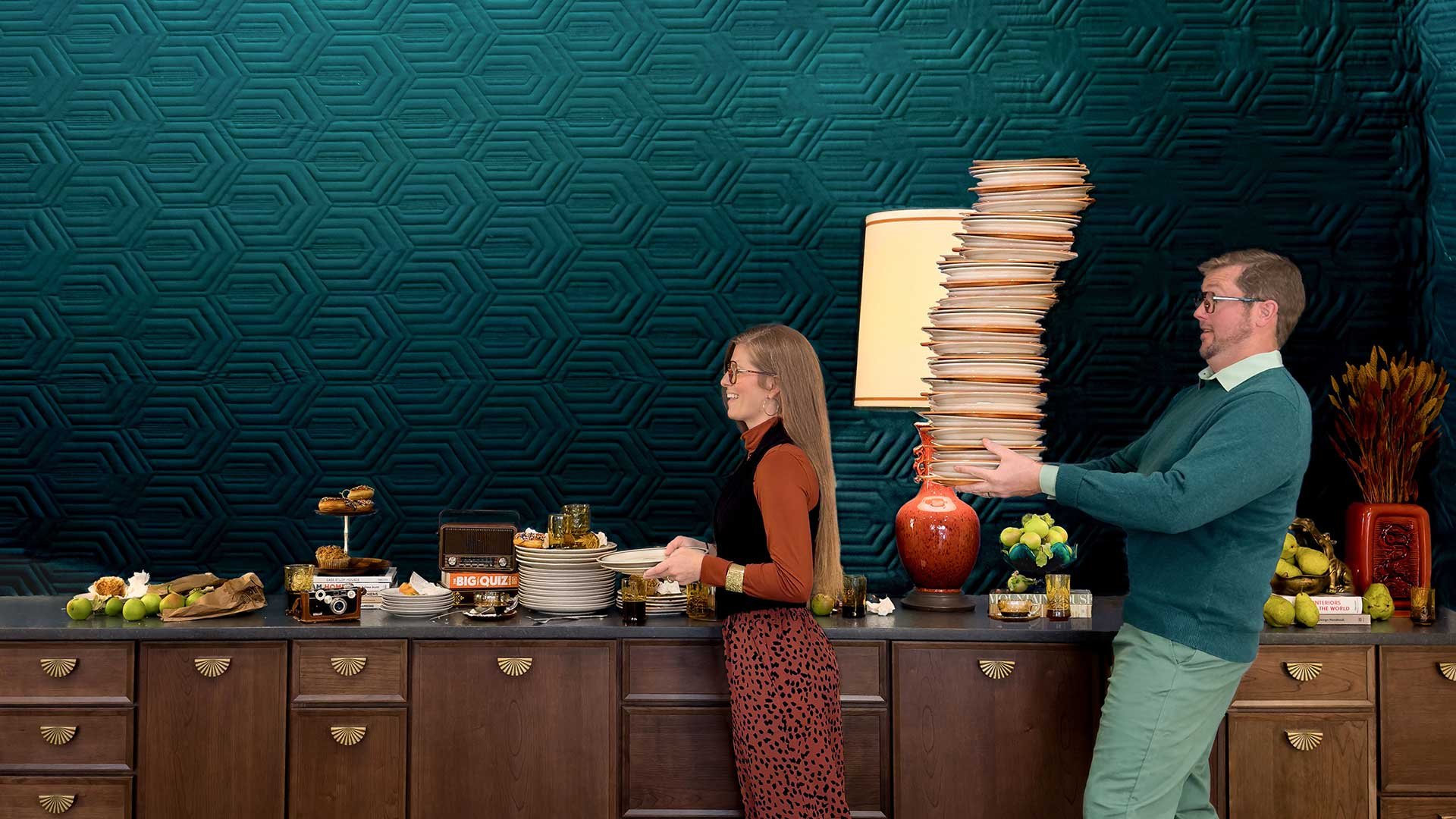 Anna carrying one dish, followed by Dane carrying a large stack of dishes. A beautifulyl designed kitchen and beautiful, modern cabinets can be seen as well.